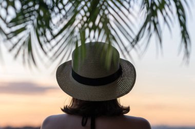 Hasır şapkalı güzel bir kızın, palmiye dallarında denizin arka planına bakan görüntüsü. Gün batımı kumsalı. Yaz ve özgürlük kavramı.