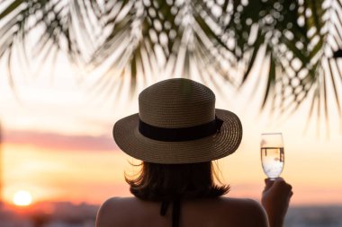 Yalnız ve güzel bir kızın arka planında hasır bir şapkayla palmiye dallarında denizin arka planına bakan bir kız. Gün batımı kumsalı. Yaz ve özgürlük kavramı.