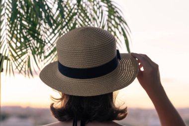 Hasır şapkalı güzel bir kızın, palmiye dallarında denizin arka planına bakan görüntüsü. Gün batımı kumsalı. Yaz ve özgürlük kavramı.