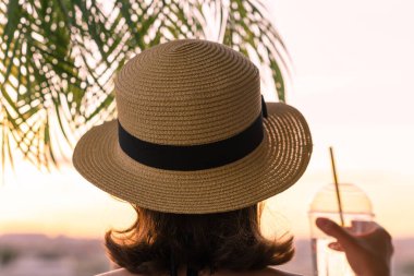 Limonlu bardakta, hasır şapkalı güzel bir kızın, palmiye dallarında denizin arka planına bakışı. Gün batımı kumsalı. Yaz ve özgürlük kavramı.