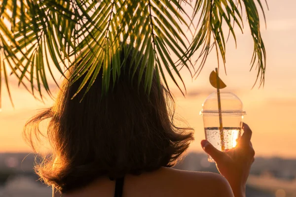 后视镜是一个漂亮的女孩 她把柠檬水放在一个塑料杯子里 背对着海的背景 在棕榈树的枝头上 日落海滩 夏天和自由的概念 — 图库照片
