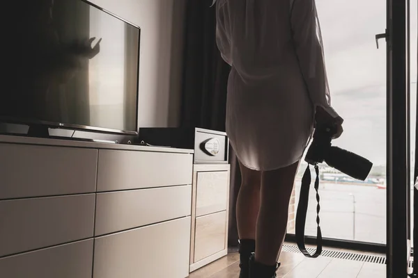 Back view of silhouette woman photographer in a transparent nightgown taking picture using camera standing near open panoramic window of apartments.