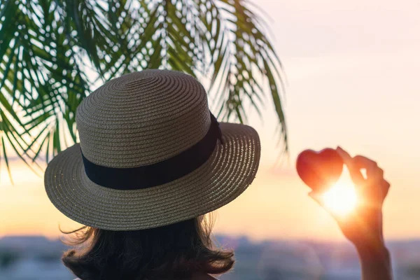 Hasır şapkalı güzel bir kızın arka planda, palmiye dallarında denizin arka planına karşı kırmızı kalbi tutuşu. Gün batımı kumsalı. Yaz ve özgürlük kavramı.