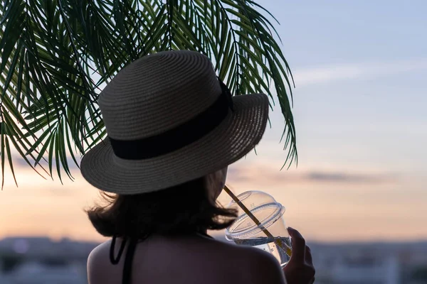 后面是一个漂亮的姑娘 戴着一顶草帽 玻璃杯里装着冷水 背对着海的棕榈树枝叶 日落海滩 夏天和自由的概念 — 图库照片