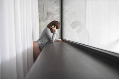 Yalnız, üzgün, beyaz bir kadın pencerenin yanında korku ve ıstırapla başını tutuyor. Depresyon, panik atak.