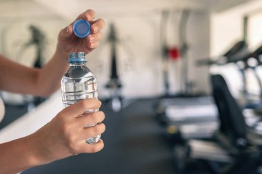 Kırpılmış. Spor malzemelerinin ve koşu simülatörlerinin karşısındaki spor salonunda elinde şişe suyu olan bir kız. Dinlenme zamanı. Sağlık kavramı.