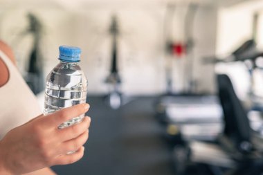 Kırpılmış. Spor malzemelerinin ve koşu simülatörlerinin karşısındaki spor salonunda elinde şişe suyu olan bir kız. Dinlenme zamanı. Sağlık kavramı.