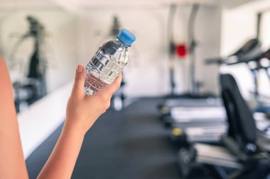 Kırpılmış. Spor malzemelerinin ve koşu simülatörlerinin karşısındaki spor salonunda elinde şişe suyu olan bir kız. Dinlenme zamanı. Sağlık kavramı.