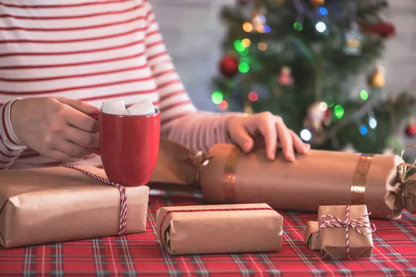 ホットチョコレートと溶融マシュマロと赤いマグカップで手作りのクリスマスプレゼントを梱包女性 輝くガーランドとボケを背景にクリスマスツリー — ストック写真