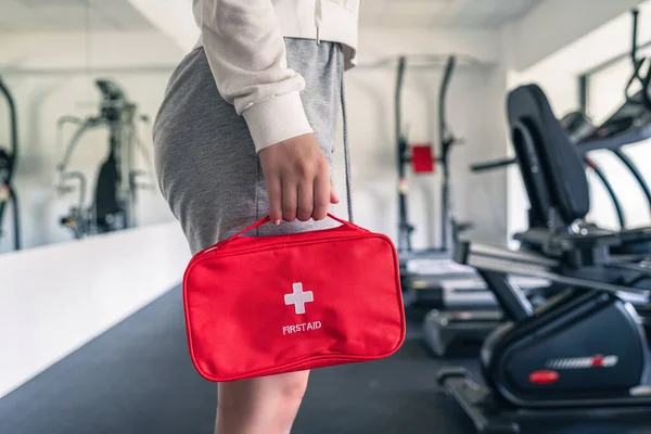 First aid kit red box in the instructor female hand in fitness gym opposite the sport equipment and  jogging simulators. Healthy lifestyle, safety and help concept.