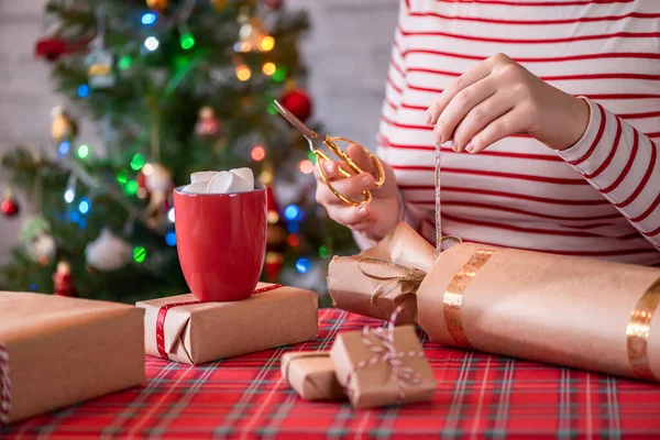 女人用红杯 热巧克力和融化的棉花糖包装手工制作的圣诞礼物 圣诞树 背景上有闪亮的花环和核桃 — 图库照片