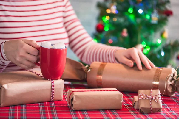 ホットチョコレートと溶融マシュマロと赤いマグカップで手作りのクリスマスプレゼントを梱包女性 輝くガーランドとボケを背景にクリスマスツリー — ストック写真