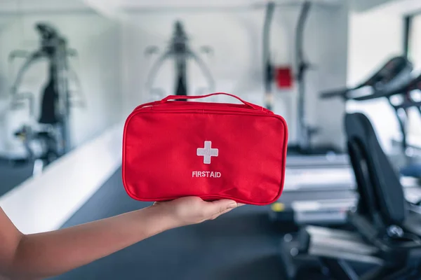 First aid kit red box in instructor female hand in fitness gym opposite the sport equipment and  jogging simulators. Healthy lifestyle, safety and help concept.