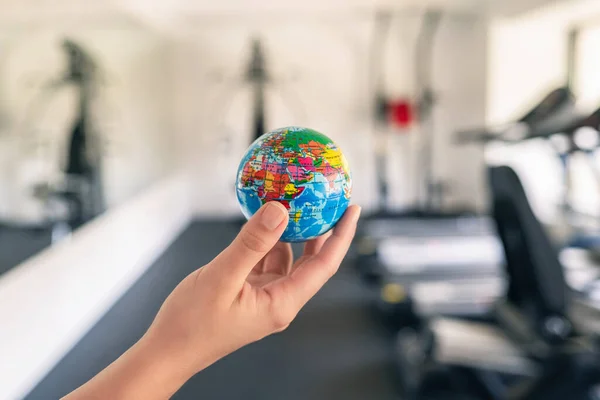 Toy globe in the female hand in fitness gym opposite the sport equipment and  jogging simulators. Healthy lifestyle. World health day. concept.