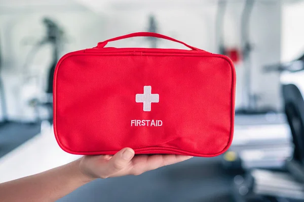 First aid kit red box in instructor female hand in fitness gym opposite the sport equipment and  jogging simulators. Healthy lifestyle, safety and help concept.