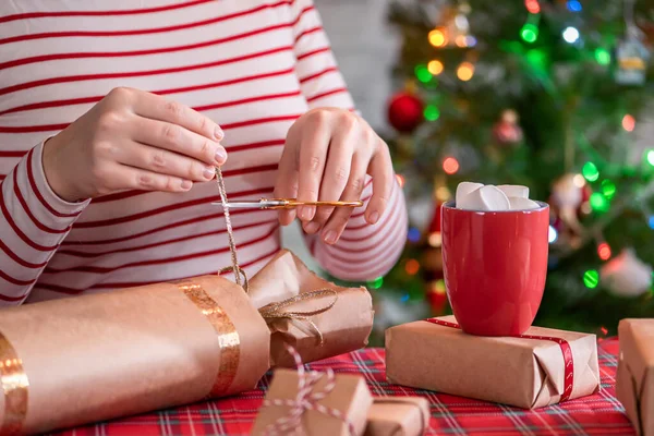 女人用红杯 热巧克力和融化的棉花糖包装手工制作的圣诞礼物 圣诞树 背景上有闪亮的花环和核桃 — 图库照片