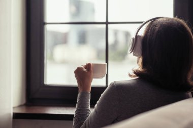 Soğuk kış sabahında pencerenin kenarında oturan, elinde kahve fincanı olan, kulaklıkla müzik dinleyen yalnız bir kadın.. 