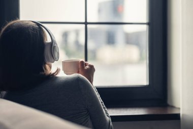 Soğuk kış sabahında pencerenin kenarında oturan, elinde kahve fincanı olan, kulaklıkla müzik dinleyen yalnız bir kadın.. 