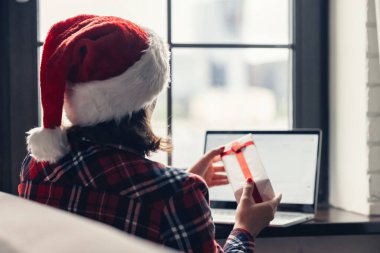 Kırmızı Noel Baba şapkalı bir kadın pencerenin yanında otururken dizüstü bilgisayar kullanıyor. Noel hediyesi alışverişi. Kavram