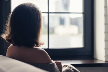 Back view of lonely aucasian young woman wrapped in a plaid enjoying sitting near window of apartments.  clipart