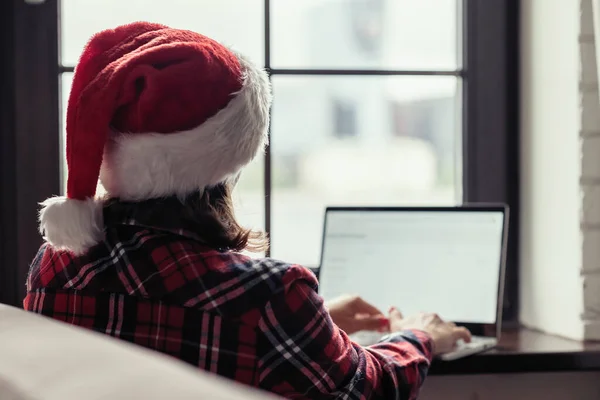 Back view of young freelancer woman in a red santa claus christmas hat sitting near window and working on laptop. Home work concept