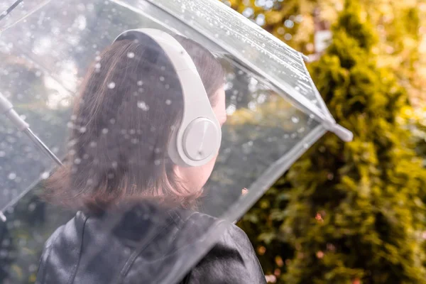庭を歩いてヘッドフォンで孤独な悲しい女性 雨滴とぬれた透明な傘を通して表示します 雨の日の風景 — ストック写真