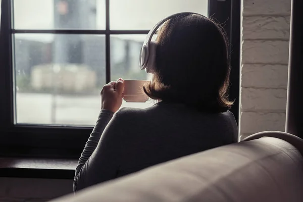 Back view of lonely aucasian young woman enjoying having breakfast with cup of coffee and listenning music in headphones sitting near window at cold winter morning.
