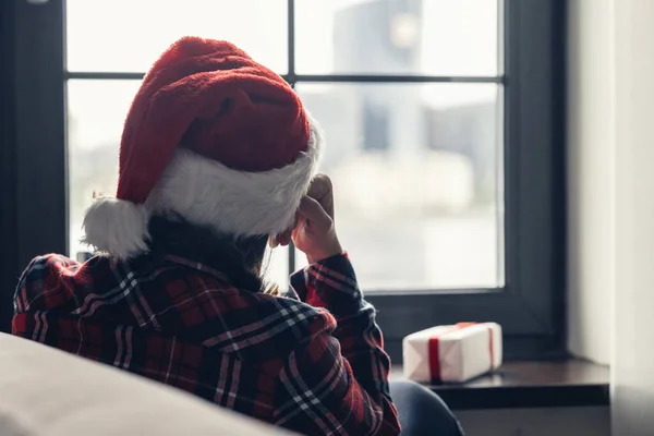赤いサンタクラスのクリスマスの帽子の孤独な悲しい若い女性のバックビューは ウィンドウの近くに座って ボーイフレンドのための贈り物を保持します コンセプト — ストック写真