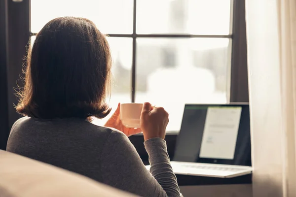 Back view of lonely woman enjoying with cup of coffee working on laptop sitting near window at morning