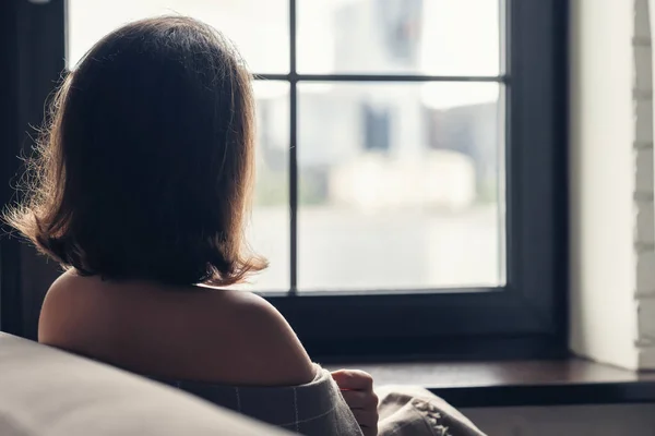 Back view of lonely aucasian young woman wrapped in a plaid enjoying sitting near window of apartments.