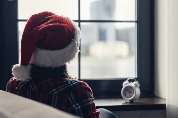 ウィンドウの近くに座って 目覚まし時計を保持赤いサンタクラスのクリスマスの帽子で孤独な悲しい若い女性のバックビュー コンセプト — ストック写真