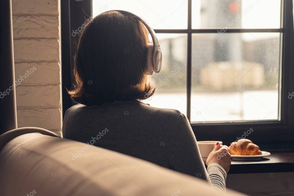 Back view of lonely aucasian young woman enjoying having breakfast with cup of hot coffee, cappuccino and croissant, listenning music in headphones sitting near window at morning. 