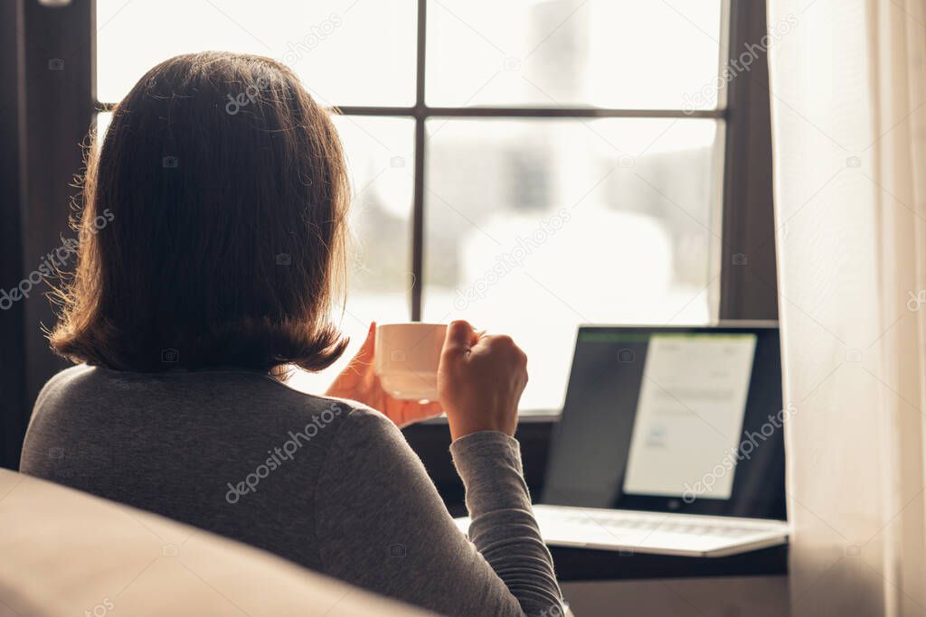 Back view of lonely woman enjoying with cup of coffee working on laptop sitting near window at morning