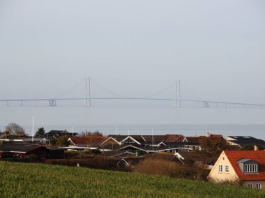 Şiddetli siste, Danimarka 'da büyük bir su kütlesinin üzerinde bir köprü. Köprü arka plandaki sisten görülebilirken, farklı evlerin çatıları ön planda görülebilir..