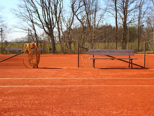 一个空旷的红土网球场的细节 中间有一条木制长椅和一条黄色的软管 — 图库照片
