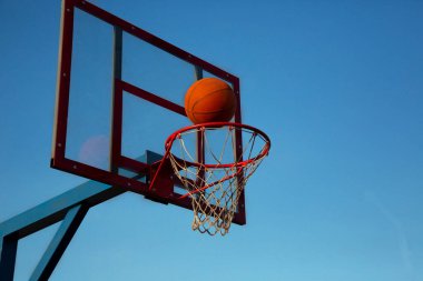 Turuncu bir basketbol topu gökyüzüne karşı açık bir alanda sepete doğru uçuyor..