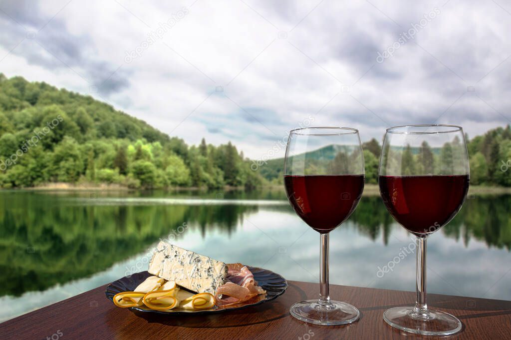 Two glasses of wine with charcuterie assortment on calm lake background in Croatia. Glass of red wine with different snacks - plate with ham, sliced, blue cheese. Romantic celebration.