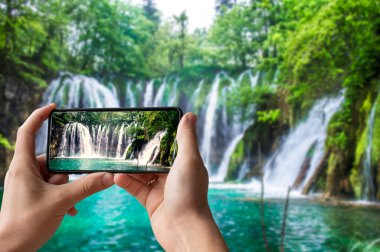 Turist zümrüt suyla şelalenin fotoğrafını çekiyor. Hırvatistan 'ın Plitvice Gölleri Ulusal Parkı' nda elinde telefon tutan ve fotoğraf çeken adam.