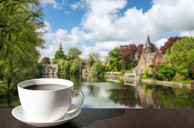Baharda güneşli bir günde, Belçika 'nın Bruges şehrinde Minnewater Gölü ve küçük bir şato manzaralı yeşil park manzaralı bir fincan kahve.