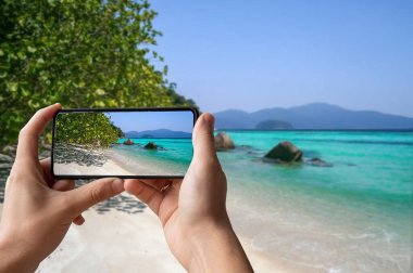 Elinde telefon tutan ve beyaz kum ve turkuaz okyanuslu tropik plajların fotoğrafını çeken bir adam. Tropikal ülkelerde tatil.