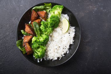 Vegan teryaki tempeh ya da tempe budha kasesi pilav, brokoli buğulama, ıspanak ve siyah arka planda limon. Sağlıklı yemek.