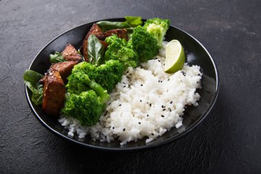 Vegan teryaki tempeh ya da tempe budha kasesi pilav, brokoli buğulama, ıspanak ve siyah arka planda limon. Sağlıklı yemek.