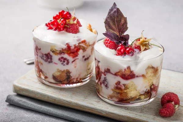 Layered trifle dessert with sponge cake, whipped cream and raspberries in serving glasses on light  background. With copy space