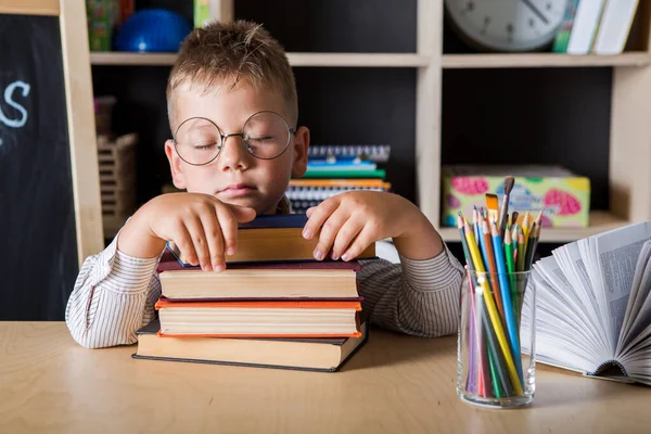 child posing while sitting at desk and lying on books in classroom. Tired boy  wearing glasses, learning process.
