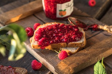 Cam kavanozda ev yapımı ahududu reçeli ve ahşap bir masada bir dilim ekmek. Yakın çekim, seçici odaklanma.