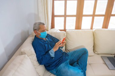 Tıbbi maskeli bir adam cep telefonuyla kanepede mesajlaşıyor.