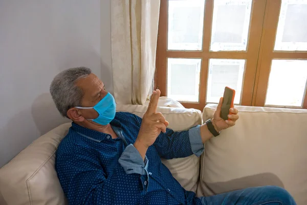 An older man in a medical mask wearing a cell phone at home, sitting on the couch, watching the screen.