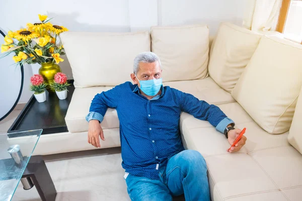 Older man with face mask who uses his cell phone on his couch at home