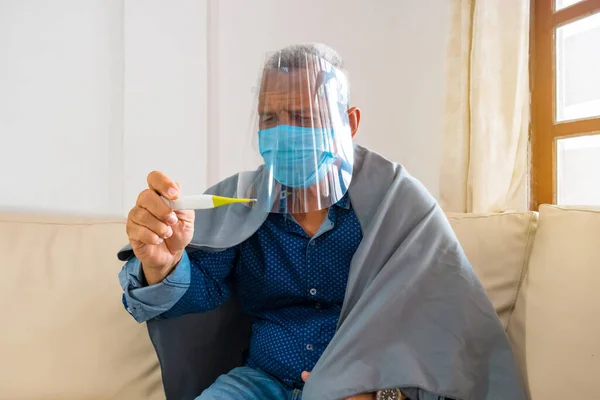 Evde termometre kullanan, kanepede oturan, kovansı 19 'lu hasta yaşlı bir adam.