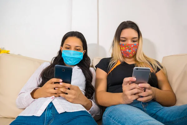 Bored friends wearing face masks and smart phones sitting on a couch in the living room